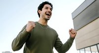 Young man with dark hair and olive skin tone wearing olive green sweatshirt celebrating success outdoors near modern building. Suitable for business, motivation, achievement, lifestyle content.