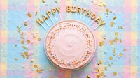 Pastel pink birthday cake with Happy Birthday candles and gold confetti on a soft plaid background, flat lay celebration concept.