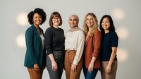 Diverse group of five women standing together against neutral background with warm lighting, wearing casual business attire. Represents teamwork, unity, and professional collaboration for corporate and diversity concepts.