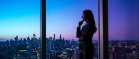 Young woman with long dark hair, wearing professional black blazer, standing by floor-to-ceiling windows in high-rise office, speaking on phone with city skyline at dusk in background. Suitable for business, corporate, and professional concepts.