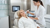 Professional dermatologist performing facial treatment on relaxed blonde woman in modern medical spa clinic with advanced equipment and sterile environment.