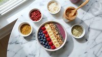 Overhead view of acai bowl with granola, fresh berries, coconut and seeds on marble surface with ingredient bowls. Food styling for healthy breakfast or smoothie bowl recipes.