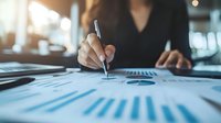 Asian businesswoman analyzing financial reports with graphs and charts at office desk, reviewing company performance data and statistics.