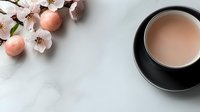 Pink cherry blossom branch with delicate petals and macarons next to black ceramic cup filled with tea on white marble surface, creating minimalist composition.