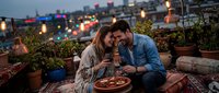 Young couple sharing a romantic rooftop dinner at dusk, laughing together with wine, surrounded by plants and warm string lights in a cozy urban setting.