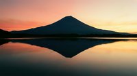Silhouetted volcanic mountain reflected in calm lake water at sunrise or sunset with warm orange and golden sky tones creating serene landscape scenery.