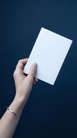 Woman's hand with silver jewelry holding blank white card against dark blue background, mockup template for text or design.