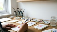 Artist painting watercolor flowers at wooden desk with brushes and palette in bright natural light studio workspace for creative hobby and art education.