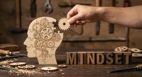 Hand placing wooden gear into brain-shaped puzzle with intricate clockwork mechanism. Mindset concept with wooden letters on rustic workshop table.