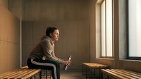 Young Asian woman sitting on wooden bench by large window holding smartphone in warm sunlit minimalist interior space for contemplation and connection.