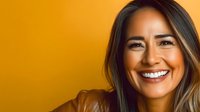 Young Asian woman with bright authentic smile wearing brown leather jacket against vibrant orange background, expressing joy and confidence in modern studio portrait.