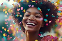 Young African American woman with natural afro hair laughing joyfully while colorful confetti falls around her, wearing red sparkly outfit against blurred background.