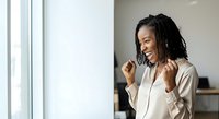 Young African American woman with long braided hair wearing a white button-up shirt, smiling while making a celebratory fist gesture in a modern office setting near a window.