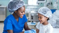 African American female nurse in blue scrubs mentoring young patient wearing protective cap in hospital setting, creating positive healthcare experience.
