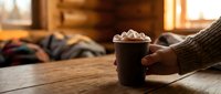 Cozy winter morning scene with hands holding hot chocolate mug topped with marshmallows in warm rustic cabin interior for seasonal comfort concepts.