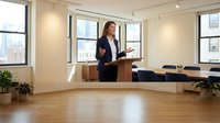 Professional businesswoman presenting in modern conference room with city view windows and wooden floors for corporate training seminars.