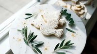 Decorated sugar cookies with intricate white icing patterns arranged on plate with fresh eucalyptus leaves for elegant dessert presentation.