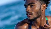 Young African American male athlete with water droplets on skin against blue ocean background, expressing vitality and wellness during outdoor workout.