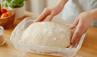 Female hands kneading fresh yeast dough on parchment paper. Homemade bread preparation process with flour dusting and wooden table background.