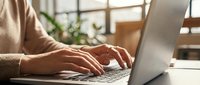 Close-up of hands typing on a laptop keyboard in a bright, modern workspace with natural light and green plants in the background.