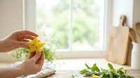 Young woman with fair skin holding a fresh yellow star fruit in bright kitchen with natural window light, wooden cutting board and fresh herbs visible, suitable for food, cooking, healthy lifestyle and culinary content.