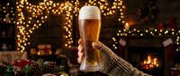 Hand holding a cold beer glass with festive holiday decorations, warm fireplace glow, and golden bokeh lights in cozy winter celebration setting.