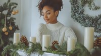 Young African American woman with natural curly hair in white knit sweater arranges Christmas decorations with white candles, pine cones and evergreen branches on table.