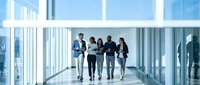 Business team walking through modern office corridor with floor-to-ceiling windows discussing project collaboration in corporate environment.