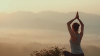 Woman practicing yoga meditation pose at sunrise in misty mountain landscape with layered hills creating peaceful wellness atmosphere for mindfulness retreat.