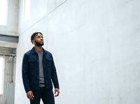 Bearded male with dark skin tone wearing navy denim jacket and gray shirt standing against white concrete wall in modern urban setting, looking upward thoughtfully.