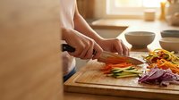 Person with fair skin chopping fresh vegetables on wooden cutting board in bright kitchen, preparing colorful bell peppers and red onions for cooking or meal preparation.