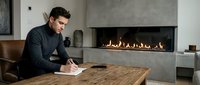 Young professional man writing in notebook at wooden table near modern fireplace in contemporary living room interior.