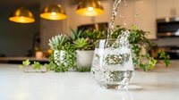 Fresh water pouring into glass on modern kitchen counter with succulent plants and warm pendant lighting creating healthy lifestyle atmosphere.