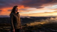 Young woman with long dark hair, Caucasian ethnicity, wearing brown jacket, standing on mountain overlooking dramatic sunset landscape with golden and orange sky, clouds, and valley below.