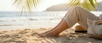 Woman relaxing on tropical beach at sunset with palm fronds overhead, wearing linen pants while sitting on sand near ocean waves for vacation lifestyle content.