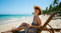 Young woman in patterned dress and sun hat relaxing on wooden beach chair by turquoise ocean with palm trees, tropical vacation lifestyle concept.