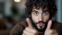 Young Middle Eastern man with curly dark hair and beard showing surprised expression with wide blue eyes and raised eyebrows while gesturing with hands.