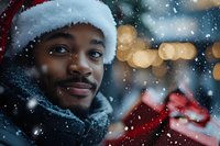 Young African American man wearing Santa hat and winter scarf smiling during snowfall with festive bokeh lights and gift box in background creating magical holiday atmosphere.