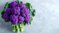 Vibrant purple cauliflower with fresh green leaves arranged on light textured background. Natural organic produce showing unique vegetable variety for healthy eating concept.