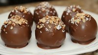 Dark chocolate truffles topped with crushed nuts on white ceramic plate, close up view of homemade confectionery dessert with smooth glossy coating.