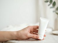 Caucasian woman's hand holding blank white cosmetic tube on neutral beige surface with soft natural lighting and blurred green plant in background, suitable for skincare and beauty product marketing.