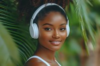 Young African American woman wearing white wireless headphones smiling confidently against green tropical foliage background, natural beauty and lifestyle portrait.