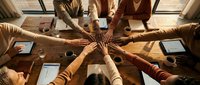 Business team joining hands together in unity gesture over wooden conference table with laptops and documents during corporate meeting.