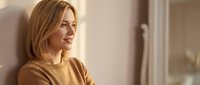 Young blonde woman in cozy sweater smiling while looking out window in warm natural light. Relaxed indoor lifestyle moment for wellness and comfort themes.