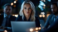 Young blonde businesswoman working late on laptop in dark office with bokeh lights and coworkers in background, professional corporate environment.