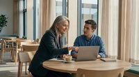 Professional mature businesswoman and young male colleague collaborating on laptop in modern office cafe during productive meeting.