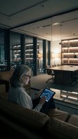 Senior man with gray hair and glasses reviewing financial data on tablet in modern office lounge overlooking illuminated workspace at night, suitable for business technology and corporate concepts.