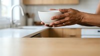 Person of African descent holding white bowl in modern kitchen with wooden countertop and stainless steel sink, suitable for food, cooking, and lifestyle content.