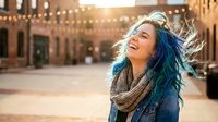 Young woman with blue hair laughing joyfully in urban plaza with string lights at sunset, expressing happiness and freedom in city environment.
