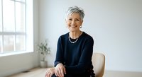 Senior woman with short gray hair wearing dark navy sweater and pearl necklace, smiling at camera while seated in bright office environment with window and plant.